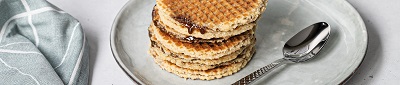 Das Bild zeigt Stroopwaffeln mit Sirup auf einem grauen Teller und daneben liegt ein Löffel und ein graues Tuch.