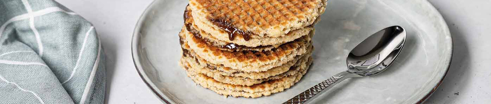 Das Bild zeigt Stroopwaffeln mit Sirup auf einem grauen Teller und daneben liegt ein Löffel und ein graues Tuch.