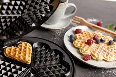 Das Bild zeigt das Waffeleisen Cloer 1898 mit einem Waffelherz, daneben steht ein Teller mit einer Herzwaffel, dekoriert mit Beeren und daneben eine graue Tasse.